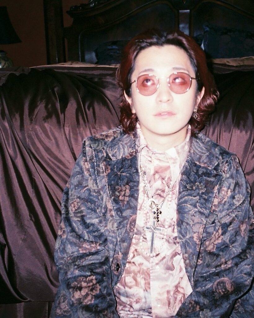 丸メガネをかけたFukaseさんが、花柄のジャケットと柄シャツを着てソファに座っている様子。