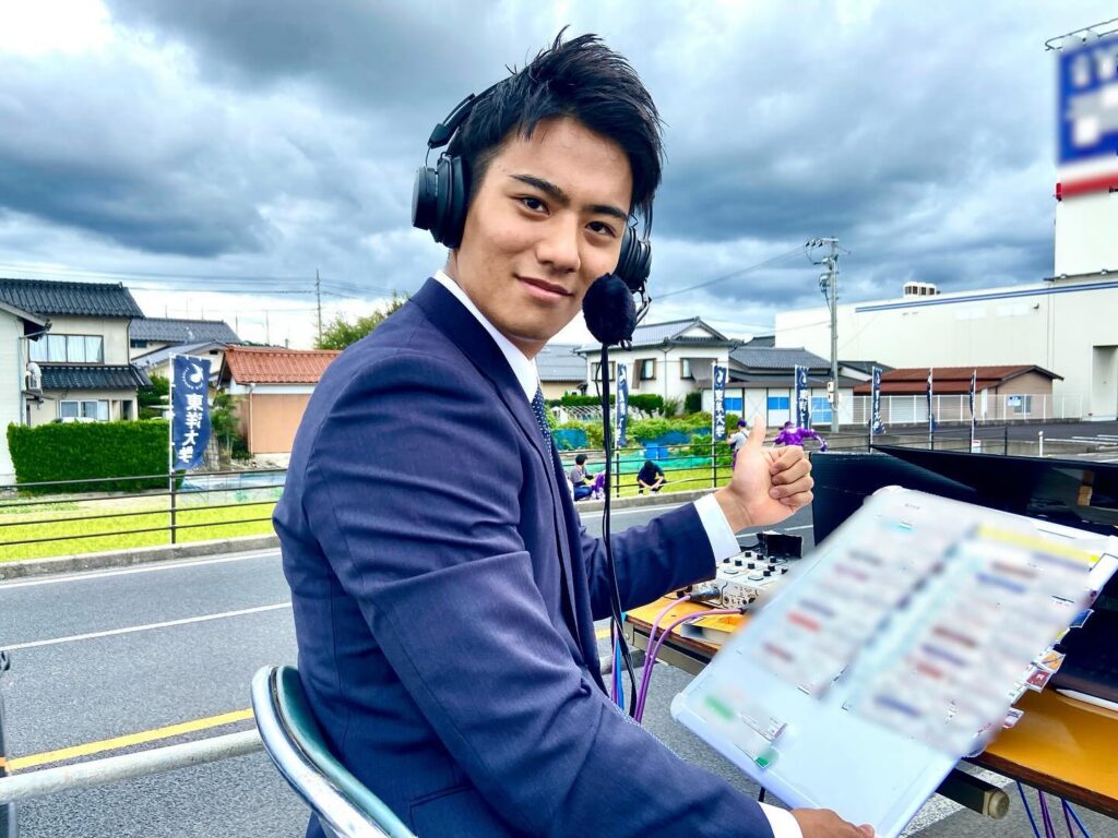 ヘッドセットを着けたスーツ姿の山本アナが、中継機材の前でポーズを取っている様子。