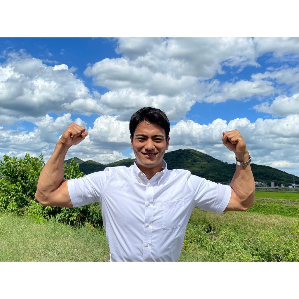 のどかな自然を背景に白いYシャツを着た山本アナが両腕を曲げて力こぶのポーズをしている様子。