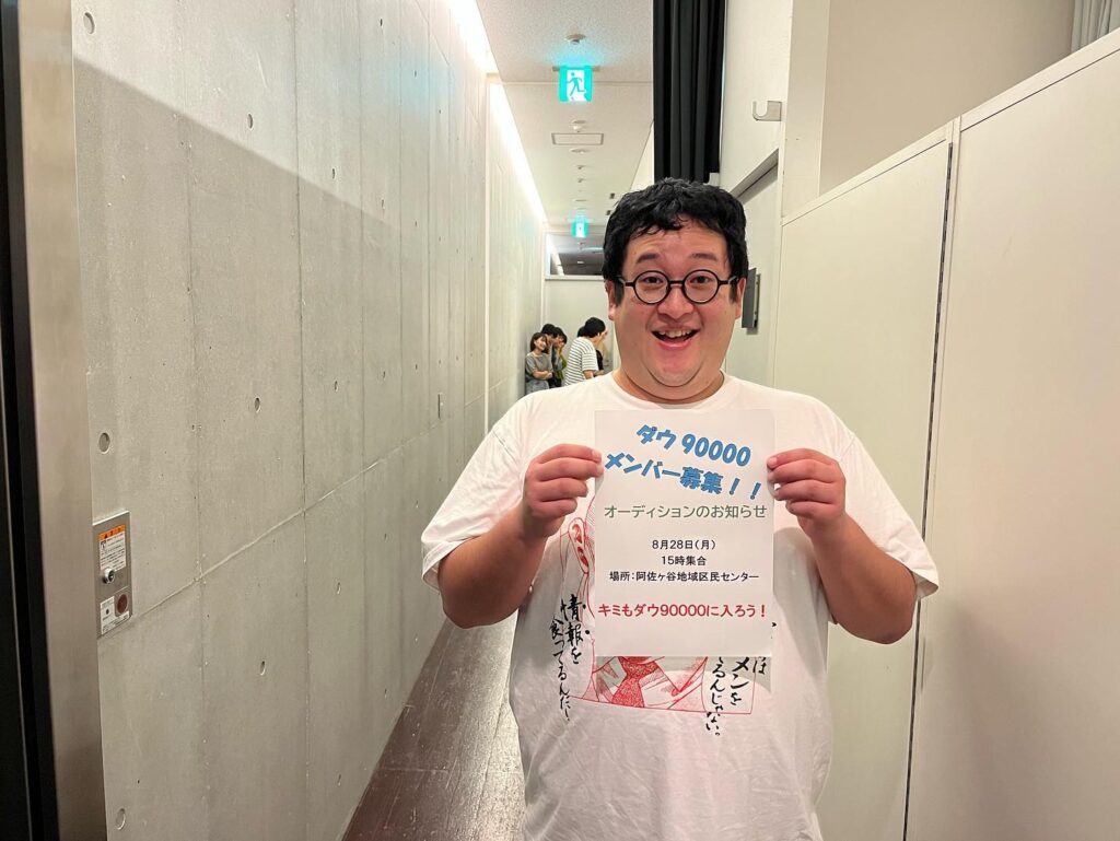 「タケ90000 メンバー募集!」と書かれた紙を持って笑顔で立つぐんぴぃさん