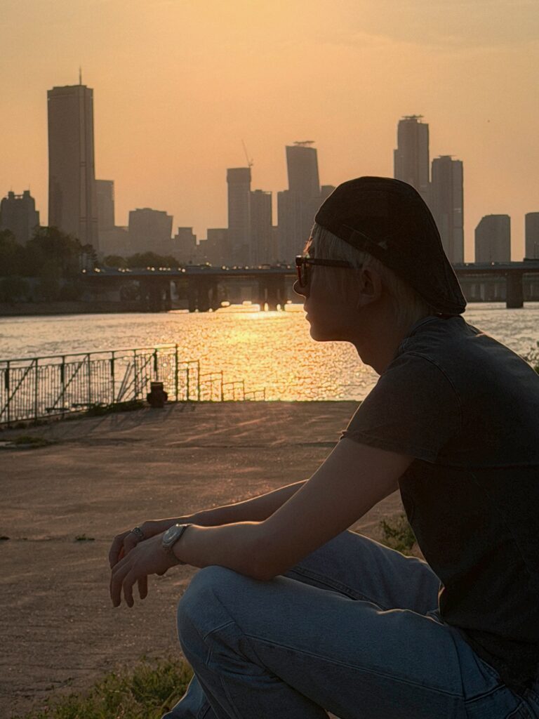 夕日を浴びる都会の川辺を背景に佇むヒスン(EVAN)さん