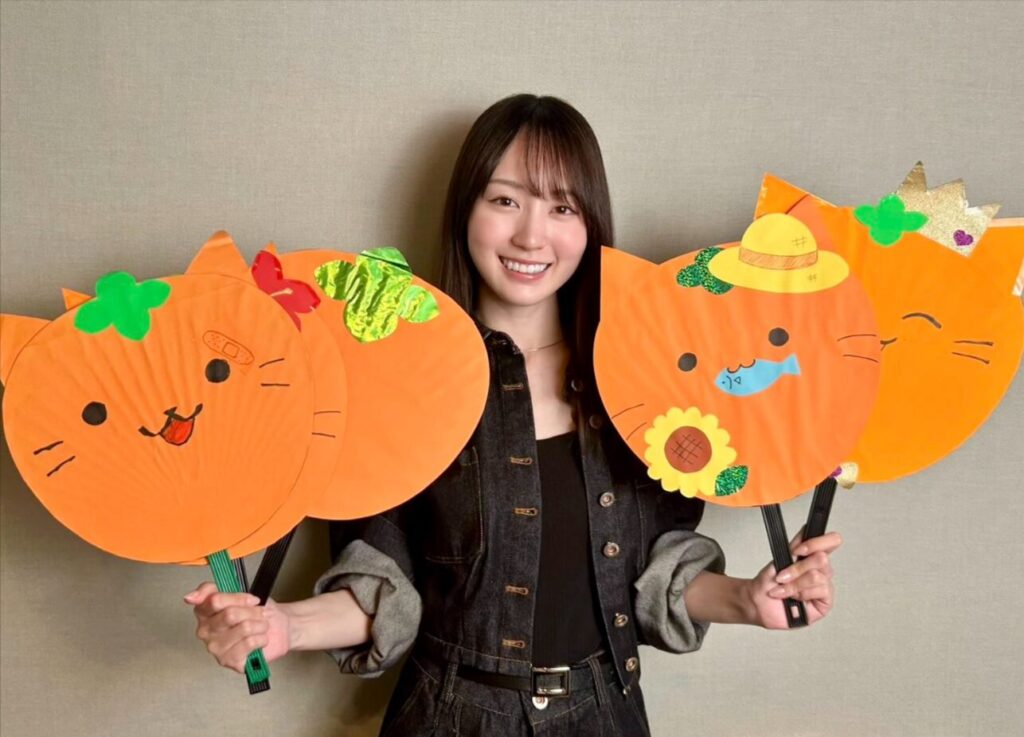 猫のイラストが描かれたオレンジ色のうちわを手に笑顔を見せる賀喜遥香さん