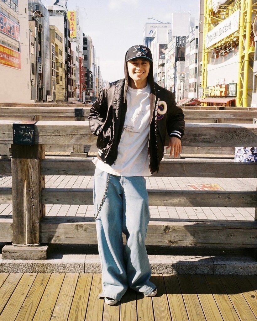 晴れた日の屋外で、木製の欄干に寄りかかりながら笑顔を見せる小島健さん