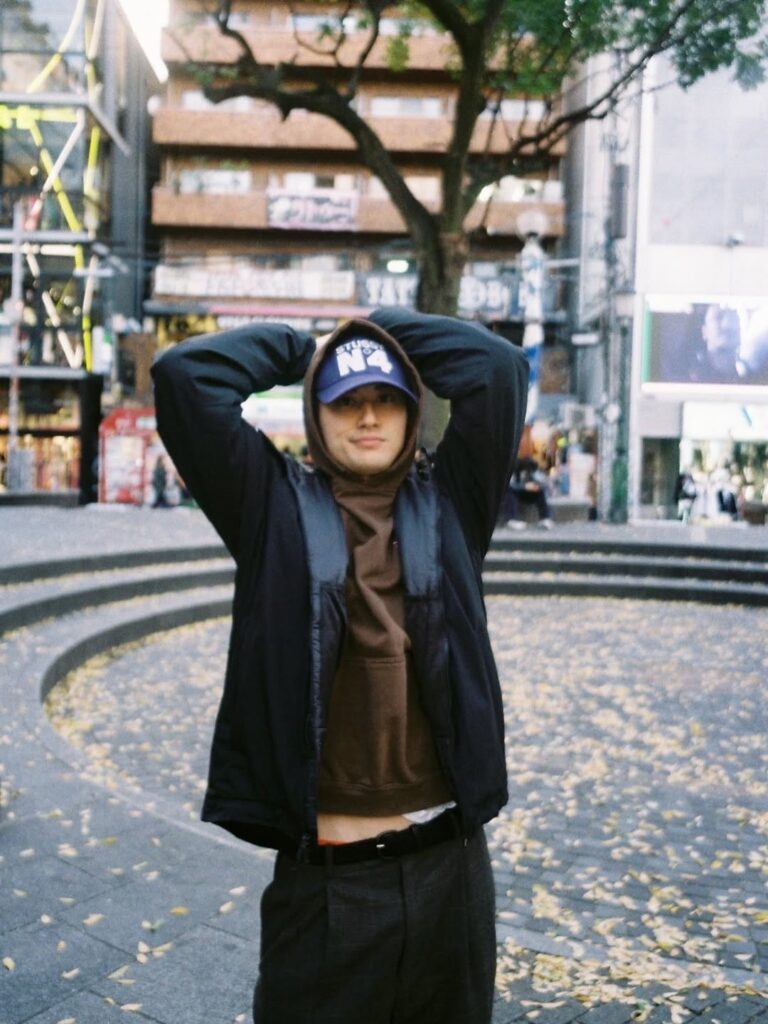 屋外の広場で、両手を頭の後ろに添えてポーズをとる小島健さん